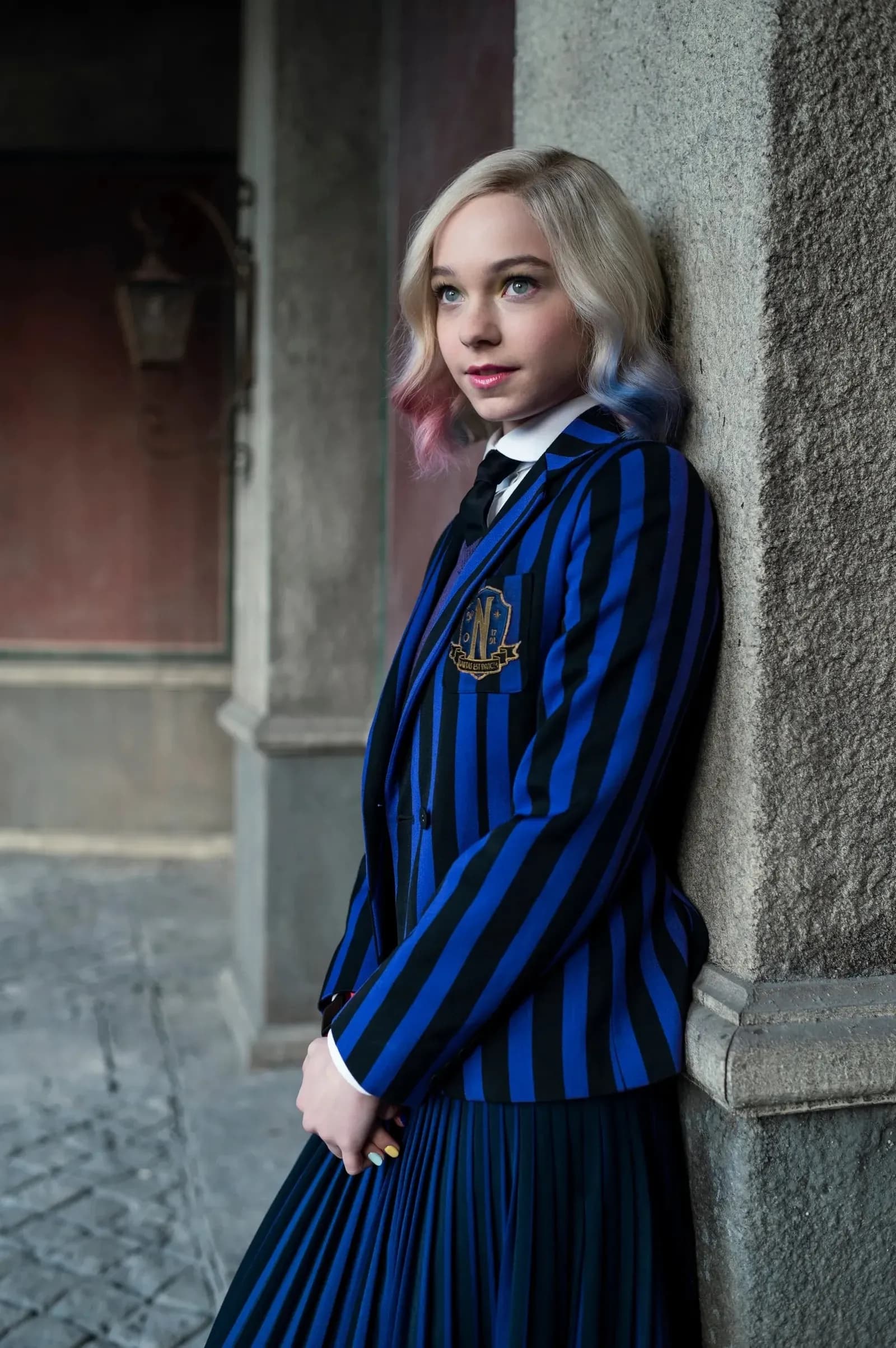 Person mit blondem, an den Spitzen rosa und blau getöntem Haar in blau-schwarz gestreiftem Blazer und Faltenrock.