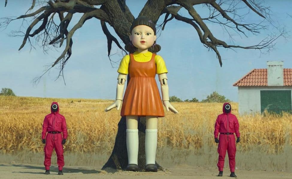 Große Puppe mit orangefarbenem Trägerkleid und gelbem Oberteil vor einem Baum.