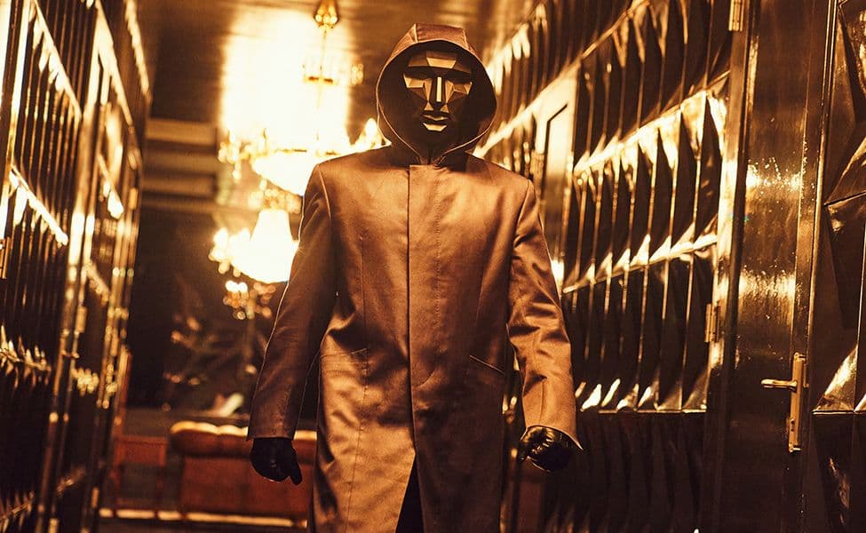Vermummte Person mit dunkler Kapuzenjacke, schwarzer kantiger Maske und Handschuhen in einem Flur.