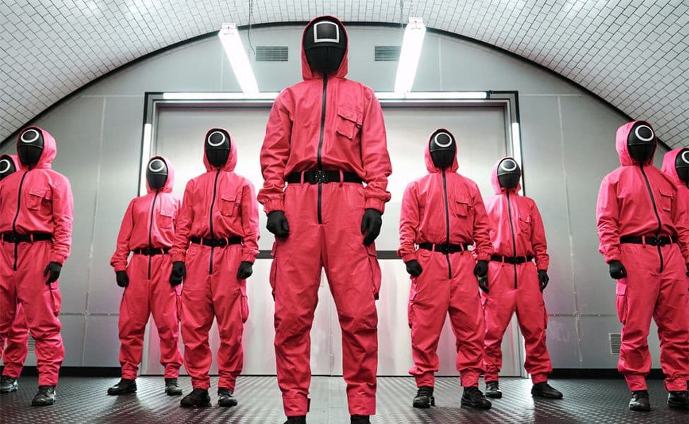 Mehrere Personen in pinkfarbenen Kapuzenoveralls mit schwarzen Masken, Gürteln, Handschuhen und Stiefeln.